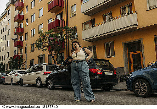 Porträt einer jungen selbstbewussten Frau  die mit der Hand auf der Hüfte vor einem Auto steht