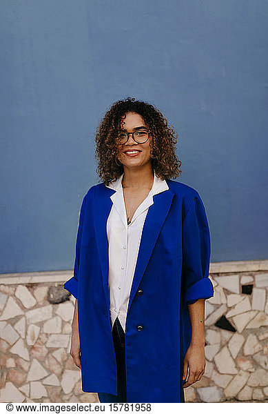 Porträt einer lächelnden Frau mit Brille und blauem Blazer  blaue Wand im Hintergrund