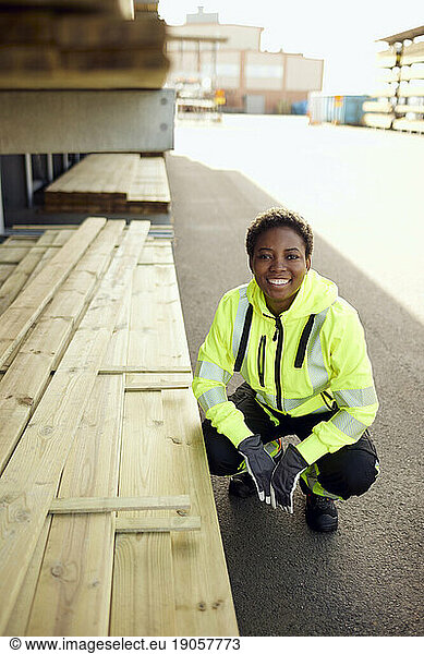 Porträt einer lächelnden jungen Arbeiterin  die an Brettern in der Holzindustrie hockt