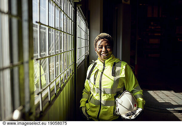 Porträt einer lächelnden jungen Arbeiterin  die in einer Fabrik am Fenster steht