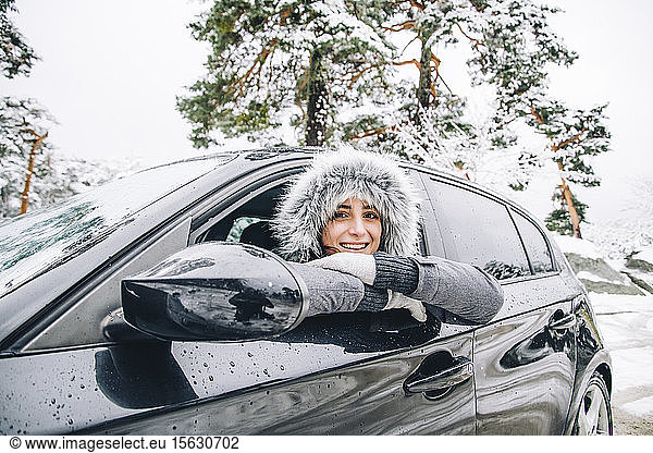 Porträt einer lächelnden jungen Frau  die sich im Winterwald aus dem Autofenster lehnt