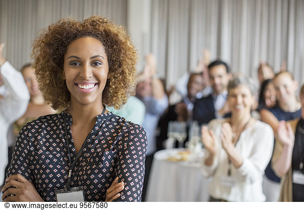 Porträt einer lächelnden jungen Frau im Konferenzraum mit applaudierenden Menschen im Hintergrund