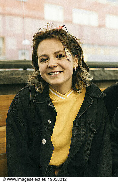 Porträt einer lächelnden jungen Frau mit Jeansjacke  die auf einer Bank sitzt