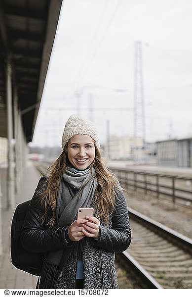 Porträt einer lächelnden jungen Frau mit Rucksack und Smartphone auf Podest stehend