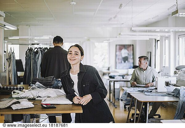 Porträt einer lächelnden jungen Modedesignerin  die sich in einer Werkstatt an eine Werkbank lehnt