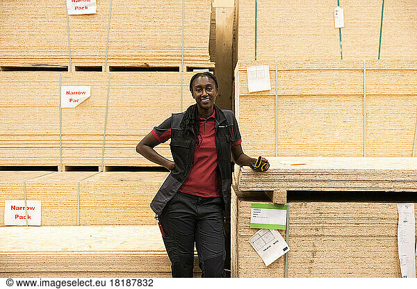 Porträt einer lächelnden Verkäuferin mit Hand auf der Hüfte  die in der Nähe eines Sperrholzstapels in einem Baumarkt steht