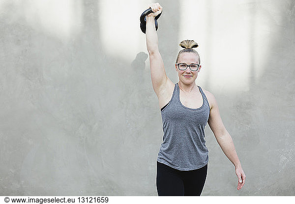 Porträt einer selbstbewussten Frau  die beim Sport die Kettlebell gegen die Wand hebt