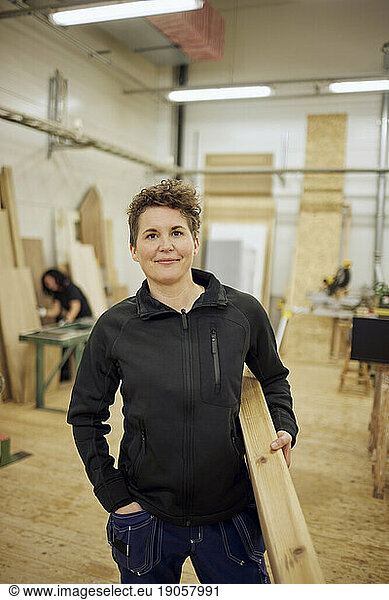 Porträt einer selbstbewussten reifen Schreinerin  die in der Werkstatt stehend eine Planke trägt