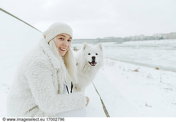 Porträt einer weiß gekleideten lächelnden jungen Frau mit ihrem weißen Hund im Schnee