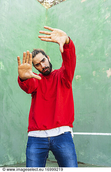 Porträt eines ernsten jungen Mannes in rotem Sweatshirt  der vor einer grünen Wand die Hände hebt