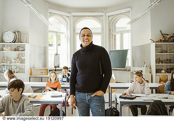 Porträt eines glücklichen männlichen Lehrers mit Hand in der Tasche  der inmitten von Schülern im Klassenzimmer steht