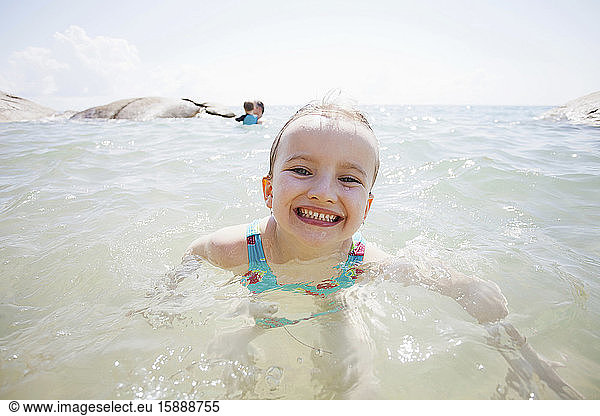 Porträt eines grinsenden kleinen Mädchens im Meer mit Vater und kleinem Bruder im Hintergrund