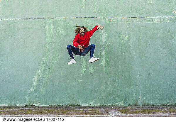 Porträt eines jungen Mannes in einem roten Sweatshirt  der vor einer grünen Wand in die Luft springt