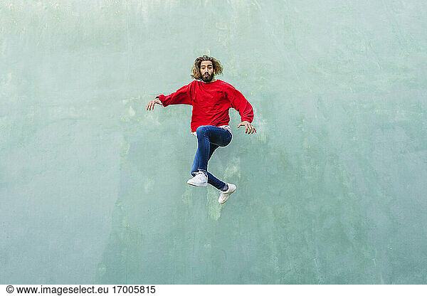 Porträt eines jungen Mannes mit rotem Sweatshirt  der vor einer grünen Wand in die Luft springt