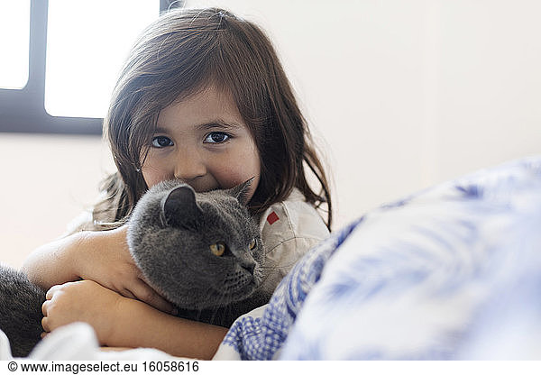 Porträt eines kleinen Mädchens  das eine graue Katze auf dem Bett kuschelt