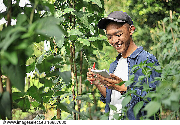 Porträt eines lächelnden jungen asiatischen Bauern  der in ein kleines Notizbuch schreibt. Glücklicher junger asiatischer Bauer im Garten