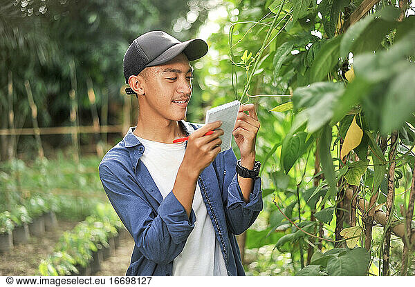 Porträt eines lächelnden jungen asiatischen Bauern  der mit einem kleinen Notizbuch die Qualität überprüft. Glücklicher junger asiatischer Bauer im Garten