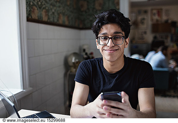 Porträt eines lächelnden jungen Mannes  der während des Studiums zu Hause soziale Medien auf seinem Mobiltelefon nutzt