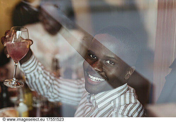 Porträt eines lächelnden jungen Mannes mit Weinglas  gesehen durch das Glas eines Restaurants