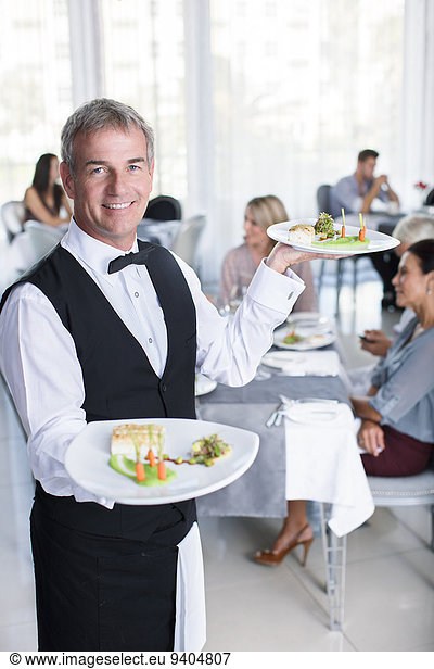Porträt eines lächelnden Kellners mit ausgefallenen Gerichten  Menschen an Restauranttischen im Hintergrund
