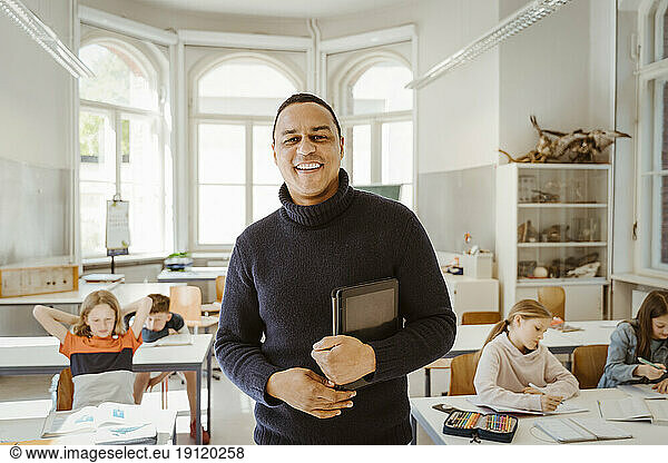 Porträt eines lächelnden männlichen Lehrers  der im Klassenzimmer stehend einen Tablet-PC hält