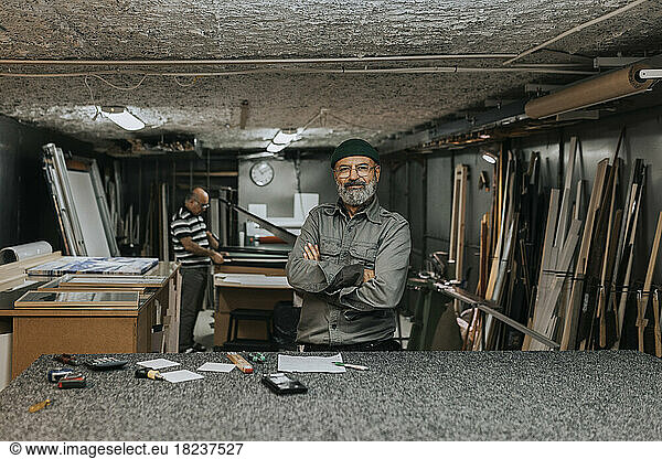 Porträt eines lächelnden männlichen Unternehmers  der mit verschränkten Armen vor einem in einer Werkstatt arbeitenden Kollegen steht