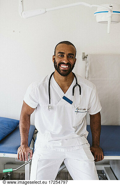 Porträt eines lächelnden medizinischen Experten  der sich auf einen Untersuchungstisch in einer medizinischen Klinik stützt