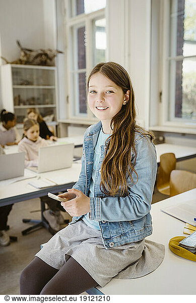 Porträt eines lächelnden Schulmädchens  das ein Smartphone in der Hand hält und am Schreibtisch im Klassenzimmer sitzt