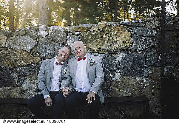 Porträt eines lächelnden schwulen Paares  das auf einer Bank vor einer Steinmauer sitzt und sich an den Händen hält