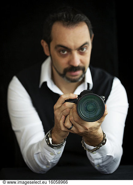 Porträt eines männlichen Fotografen  der mit seiner Kamera vor einem schwarzen Hintergrund steht  Fokus auf die Kamera im Vordergrund; Studio
