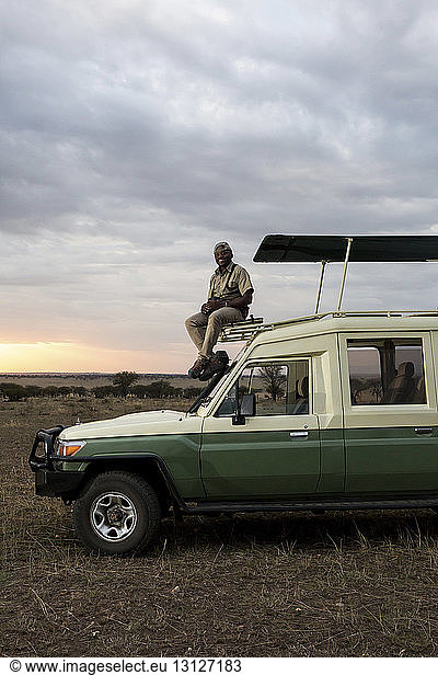 Porträt eines Mannes,  der auf einem Geländewagen vor bewölktem Himmel im Serengeti-Nationalpark sitzt