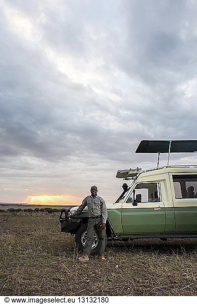 Porträt eines Mannes  der im Serengeti-Nationalpark vor bewölktem Himmel neben einem Geländewagen steht