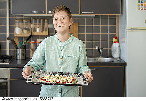 Porträt eines stolzen Jungen  der ein Backblech mit roher hausgemachter Pizza hält