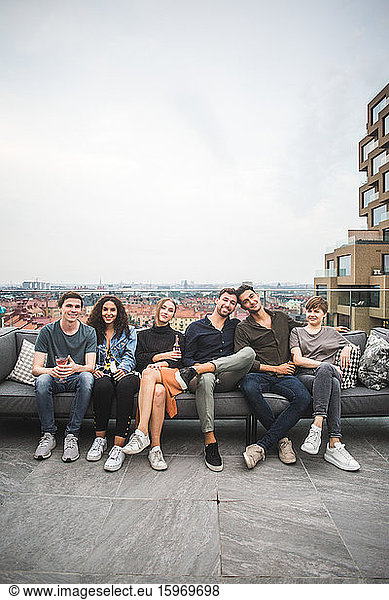 Porträt von lächelnden Freundinnen und Freunden  die auf einem Sofa auf einer Gebäudeterrasse für geselliges Beisammensein sitzen