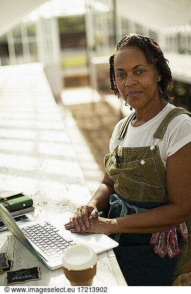 Portrait confident female garden shop owner at laptop in greenhouse