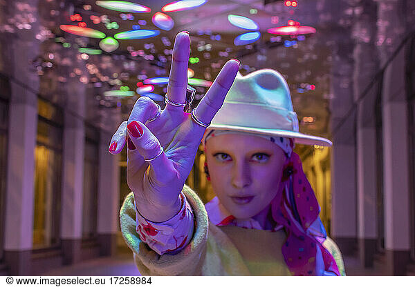 Portrait cool stylish woman in fedora gesturing peace sign under neon