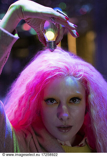 Portrait cool woman with pink hair holding light bulb over head