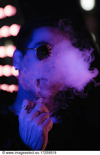 Portrait cool young man with vape pen blowing smoke in neon light