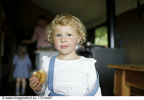 Portrait cute girl with curly blonde hair eating biscuit at home
