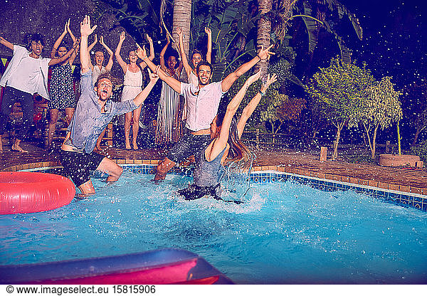 Portrait exuberant young friends jumping into swimming pool at night