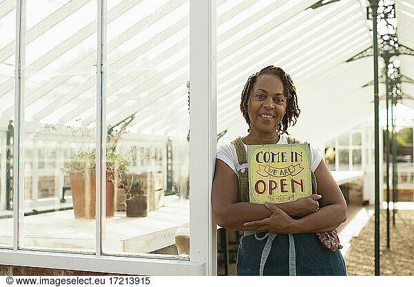 Portrait female garden shop owner with open sign in greenhouse
