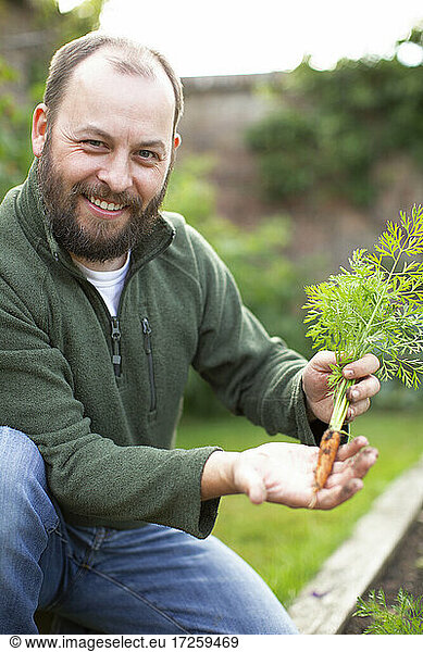 Portrait happy man with beard showing fresh harvested carrot in garden