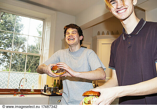 Portrait happy teenage boys eating hamburgers in kitchen
