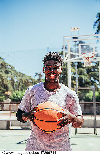 Portrait of a black African-American boy smiling and holding a basketball against his chest on an urban basketball court.