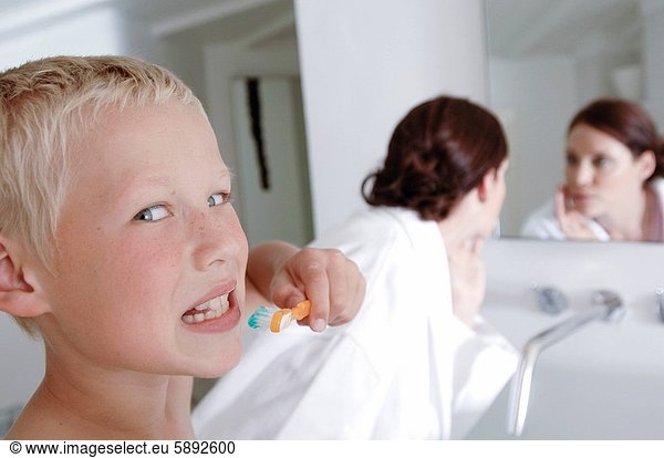 Portrait of a boy brushing teeth and his mother looking at mirror