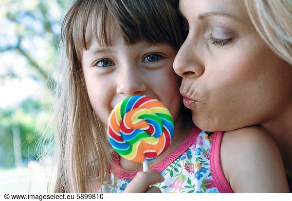 Portrait of a girl holding a lollipop with her mother kissing on her cheek