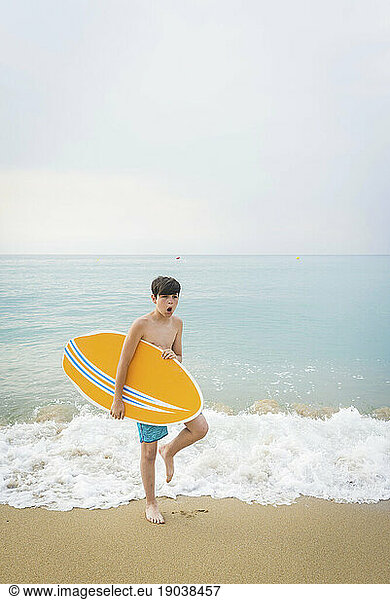 Portrait of a joyful smiling teen boy with yellow surfboard standing on the beach