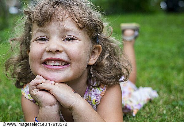 Portrait of a preschooler girl with blond curly hair and brown eyes  lying on the grass with hands folded in front of her; Toronto  Ontario  Canada