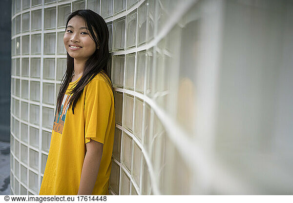 Portrait of a preteen girl wearing a yellow t-shirt and standing against a wall of glass blocks; Hong Kong  China