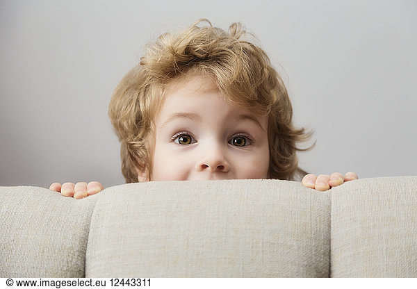 Portrait of a young boy peeking out from behind a couch and looking at the camera; Langley  British Columbia  Canada
